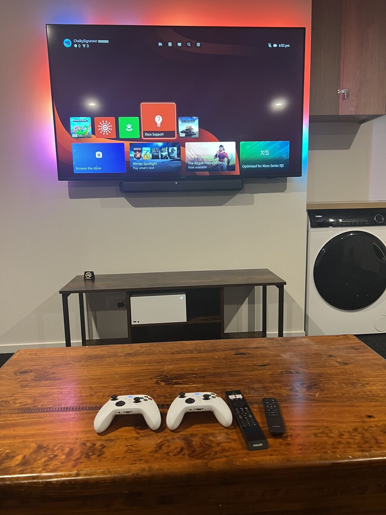 Una TV a schermo piatto montata a parete mostra varie icone di gioco e una sveglia digitale. Sotto la TV, un tavolo di legno con due controller di gioco bianchi e un telecomando.