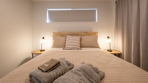 Ein ordentlich bezogenes Bett mit Kopfteil, zwei Nachttischlampen und einem Fenster mit Jalousien.