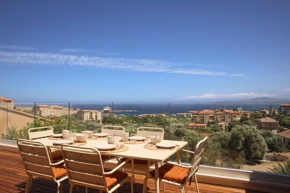 A terrace with a table set for dining, overlooking a coastal town with buildings and olive trees.