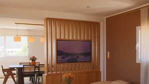 A modern living room with a large flat-screen TV mounted on a wooden panel wall, a beige sofa, a dining area with a wooden table and chairs, and a vase with flowers on a side table. - Luxury apartment for 6 people, sea view, 800m from center and beaches (L'Île-Rousse)