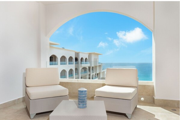 Un balcón con dos sillas acolchadas y una mesa, con vistas a un edificio con varios balcones y al mar.
