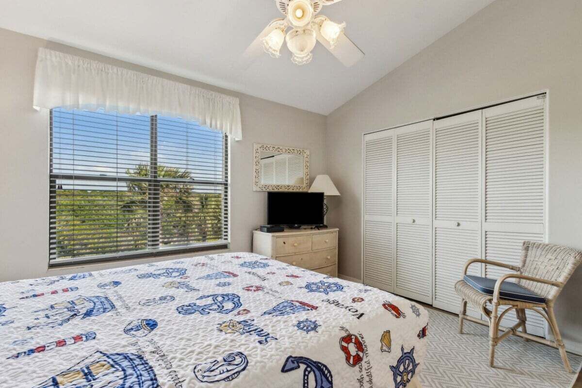 Un dormitorio con cama, una silla, televisor, ventilador de techo, ventana con persianas y armario.