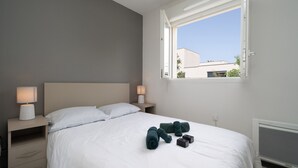 A neatly made bed with white linens, two bedside lamps, and a window with an open view.