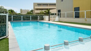 A swimming pool with a white fence, a grassy area, and a building with balconies in the background.