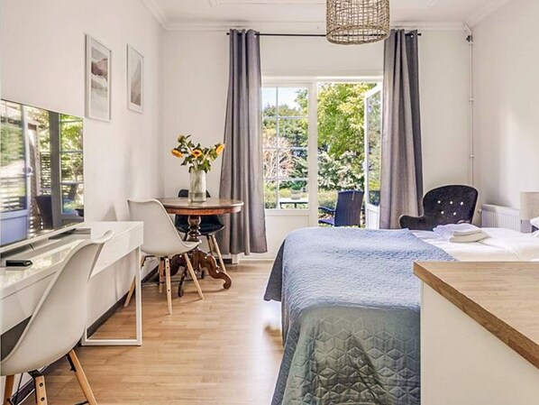 A modern bedroom with a bed, a desk, and a chair. There is a window with curtains and a view of greenery outside.