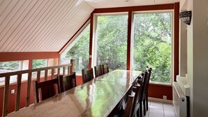 A dining area with a long table and chairs, large windows with a view of trees, and a sloped ceiling. - Group Home in La Roche With Sauna (La Roche-en-Ardenne)