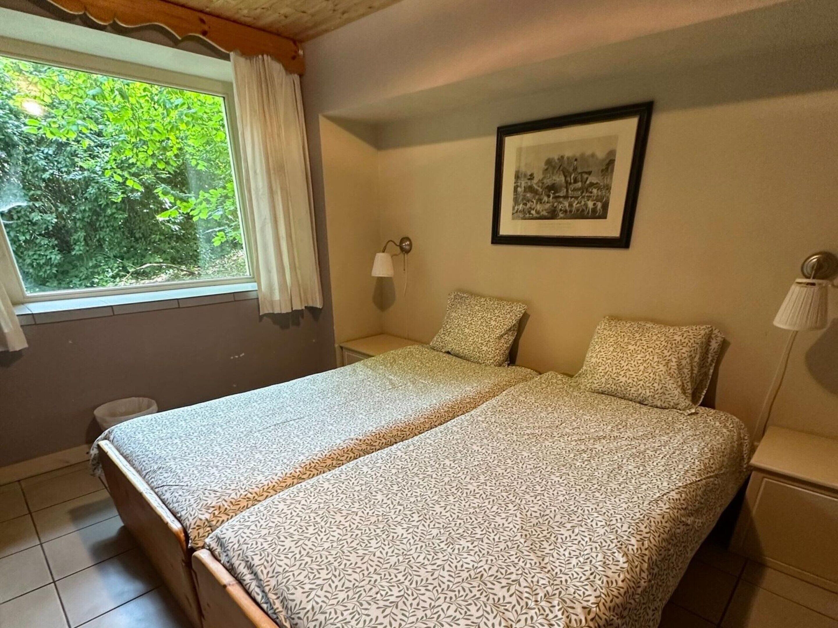A double bed with patterned bedding, a bedside table, a nightstand, a wall-mounted lamp, a framed picture, and a window with a view of greenery.
