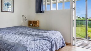 A bedroom with a bed, a bedside table, a clock, a window with curtains, and a view of greenery outside.