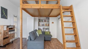 Een loft-achtige kamer met zichtbare houten balken, een ingebouwde boekenkast, een grijze bank en een houten trap.