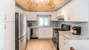 A modern kitchen with white cabinets, stainless steel appliances, a wooden ceiling, and a window. - Wasaga Beach Summer Cottages & Cabins (Wasaga Beach)