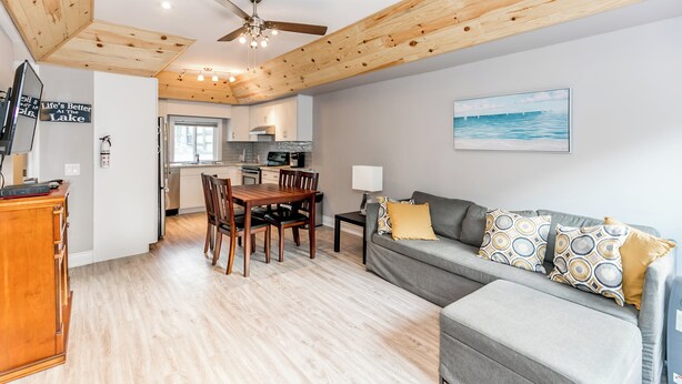 A modern kitchen with wooden ceiling beams, a dining area with a wooden table and chairs, a grey sofa with patterned cushions, and a painting of a seascape on the wall. - Wasaga Beach Summer Cottages & Cabins (Wasaga Beach)