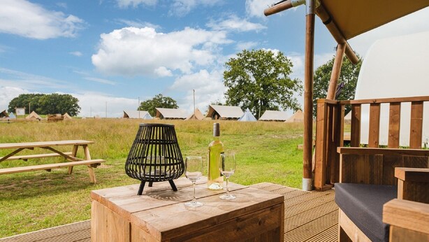 Een houten terras met een tafel, een fles en glazen, met uitzicht op een veld met tenten.