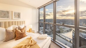 A modern bedroom with a large window overlooking a cityscape, a bed with multiple pillows, and a nightstand with a lamp.