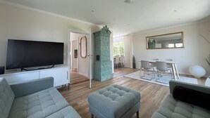 Ein Wohnzimmer mit einem Flachbildfernseher, grauen Sofas, einem türkisblauen Schrank und einem Essbereich mit weißem Tisch und Stühlen.