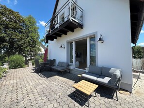 Um pátio moderno ao ar livre com piso de azulejos hexagonais, uma mesa de madeira, um sofá cinza e uma varanda com grades de metal.