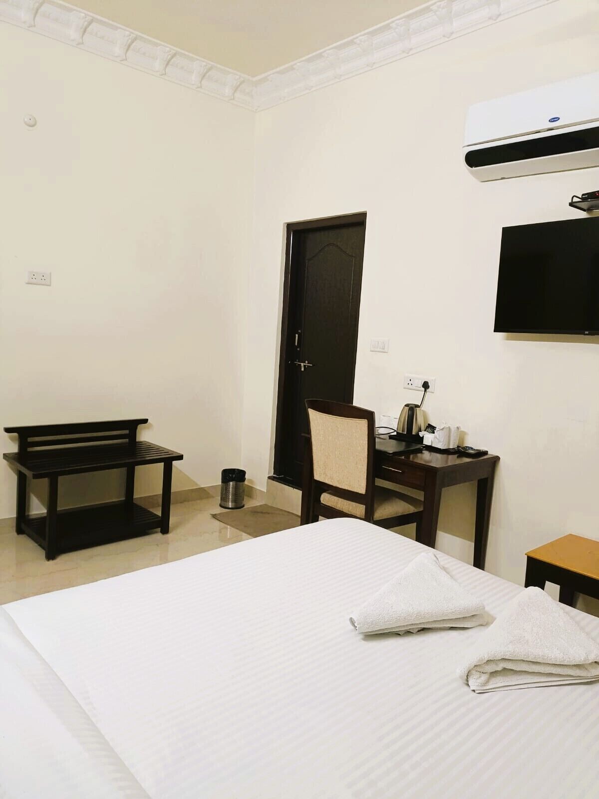 A hotel room with a bed, a desk, a chair, a bench, and an air conditioning unit.