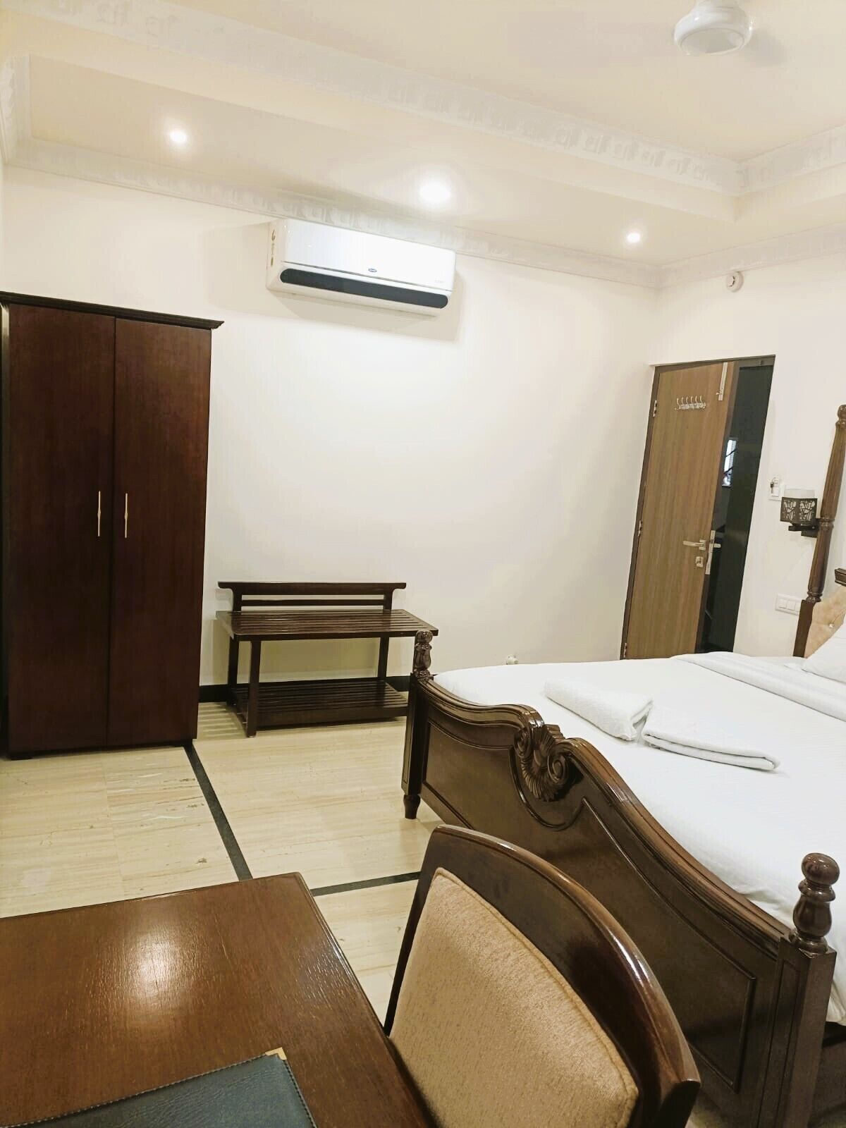 A hotel room with a wooden bed, a wooden wardrobe, a small wooden table, and a bench.