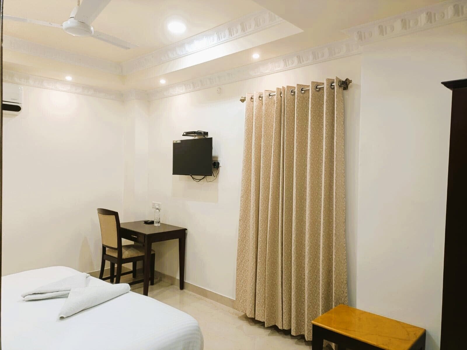 A hotel room with a bed, a desk with a chair, a television mounted on the wall, and a curtain.