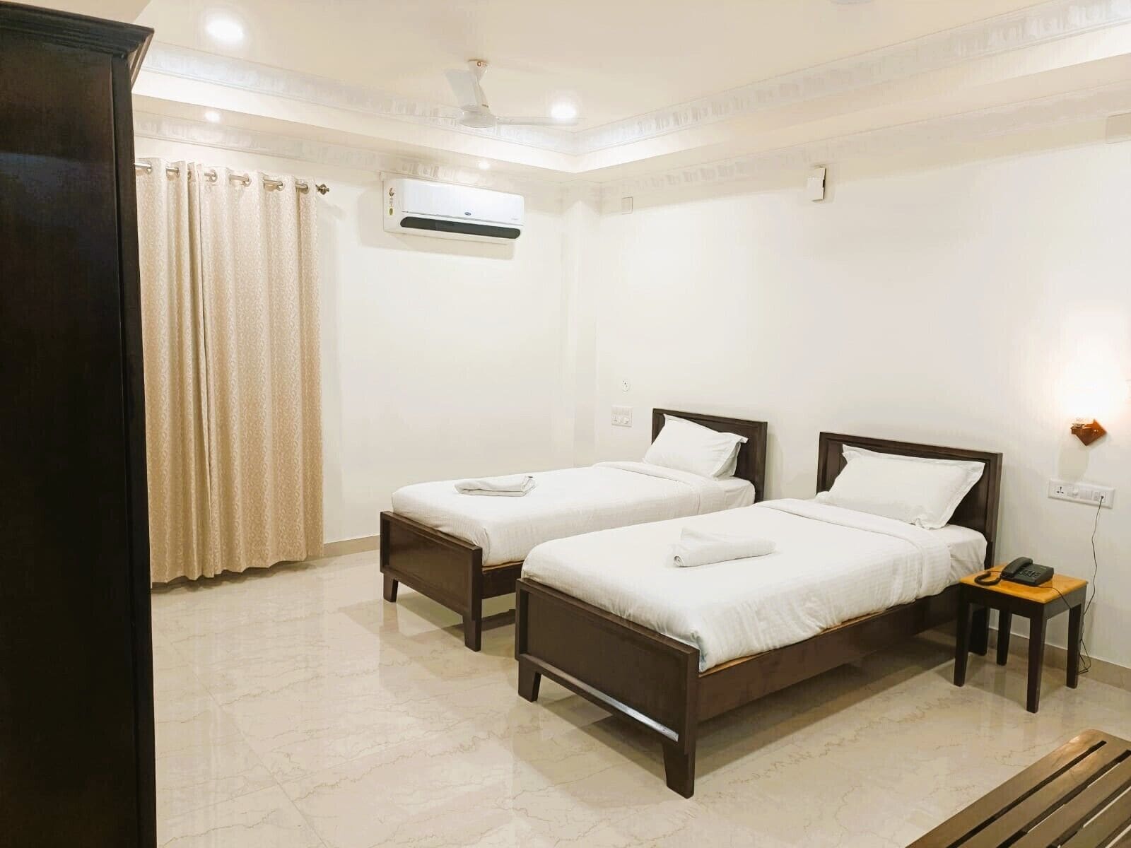 Une chambre d’hôtel avec deux lits simples, une table de chevet avec un téléphone, un climatiseur fixé au mur et un rideau.