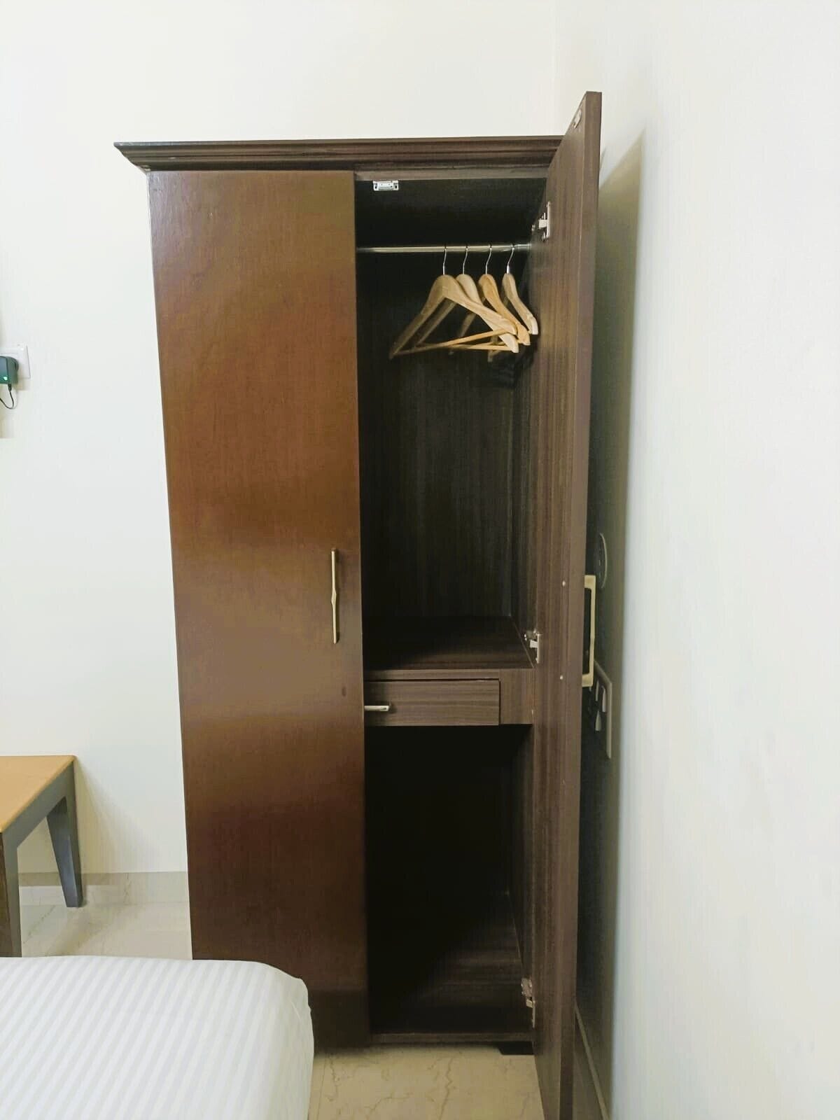 A wooden wardrobe with a hanging space and a small drawer, located in a room with a bed and a nightstand.