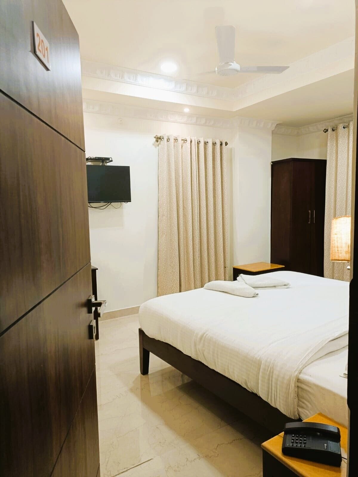 Habitación de hotel con cama, armario, televisor, cortina y mesita de noche con lámpara.