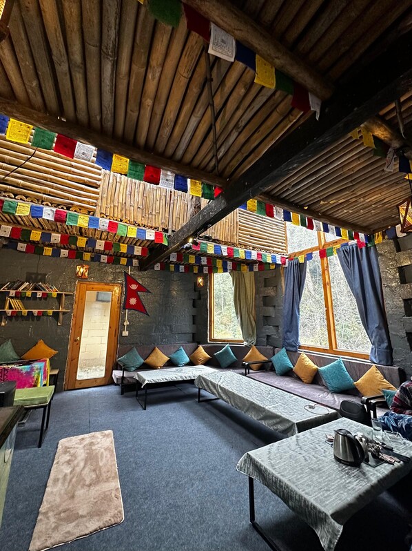 A room with a wooden ceiling, prayer flags, a sofa with cushions, a table with a kettle, and a window with curtains. - Sunshine Cafe & Cottage Riverside (Ghiyagi)