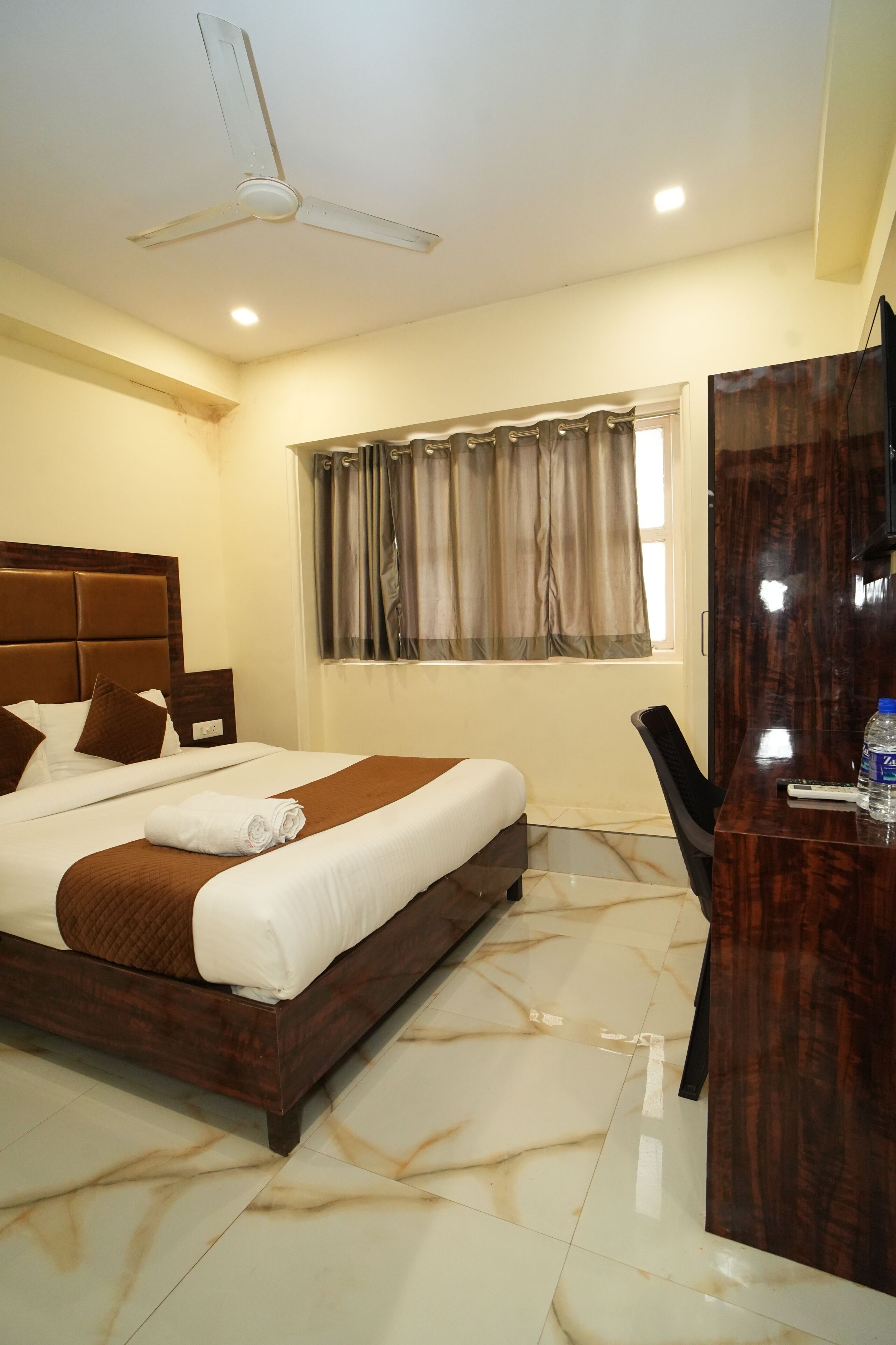Habitación de hotel con cama, escritorio, silla, televisor y ventana con cortinas.