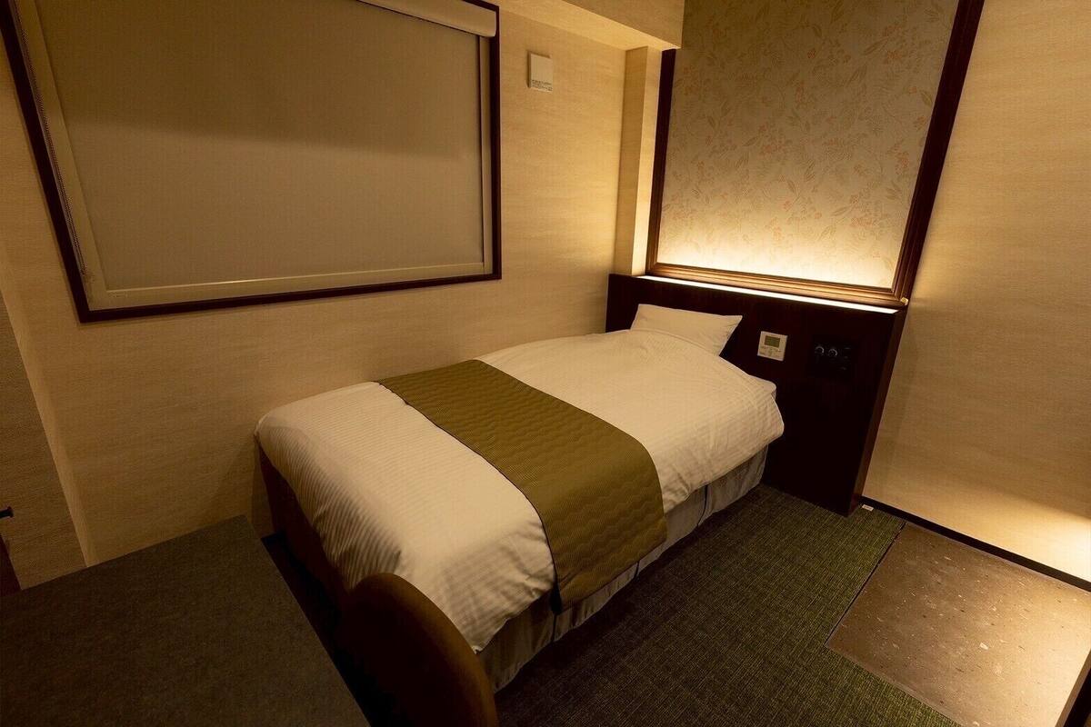 Una cama individual con una colcha verde, cabecera con luz y una mesita de noche pequeña.