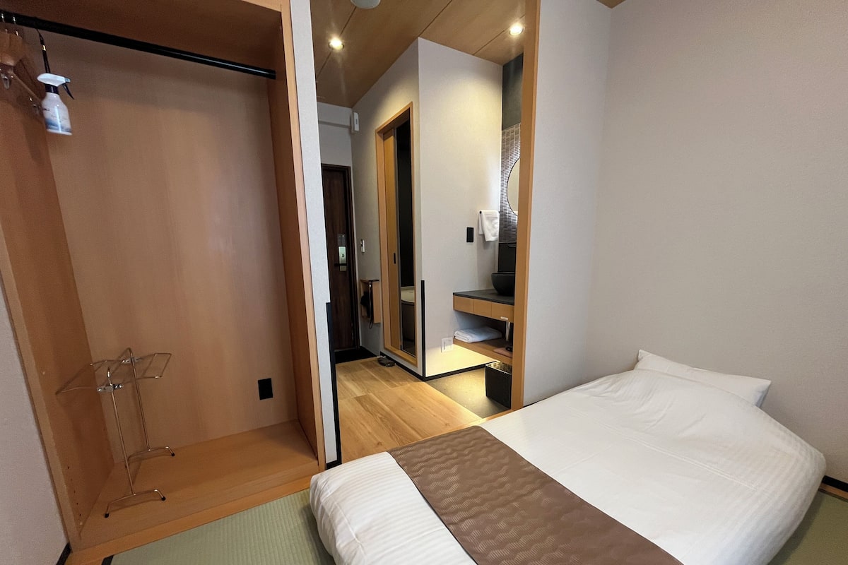 Habitación de hotel con cama, suelo de madera, baño con espejo y lavamanos, y pasillo que conduce a otra habitación.