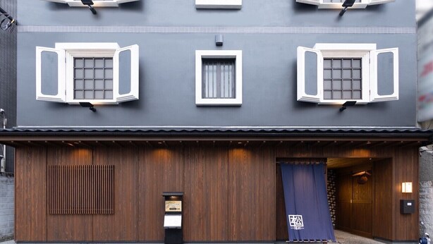 Un edificio moderno con fachada gris, varios ventanales con marcos blancos, una entrada de madera y una puerta azul.