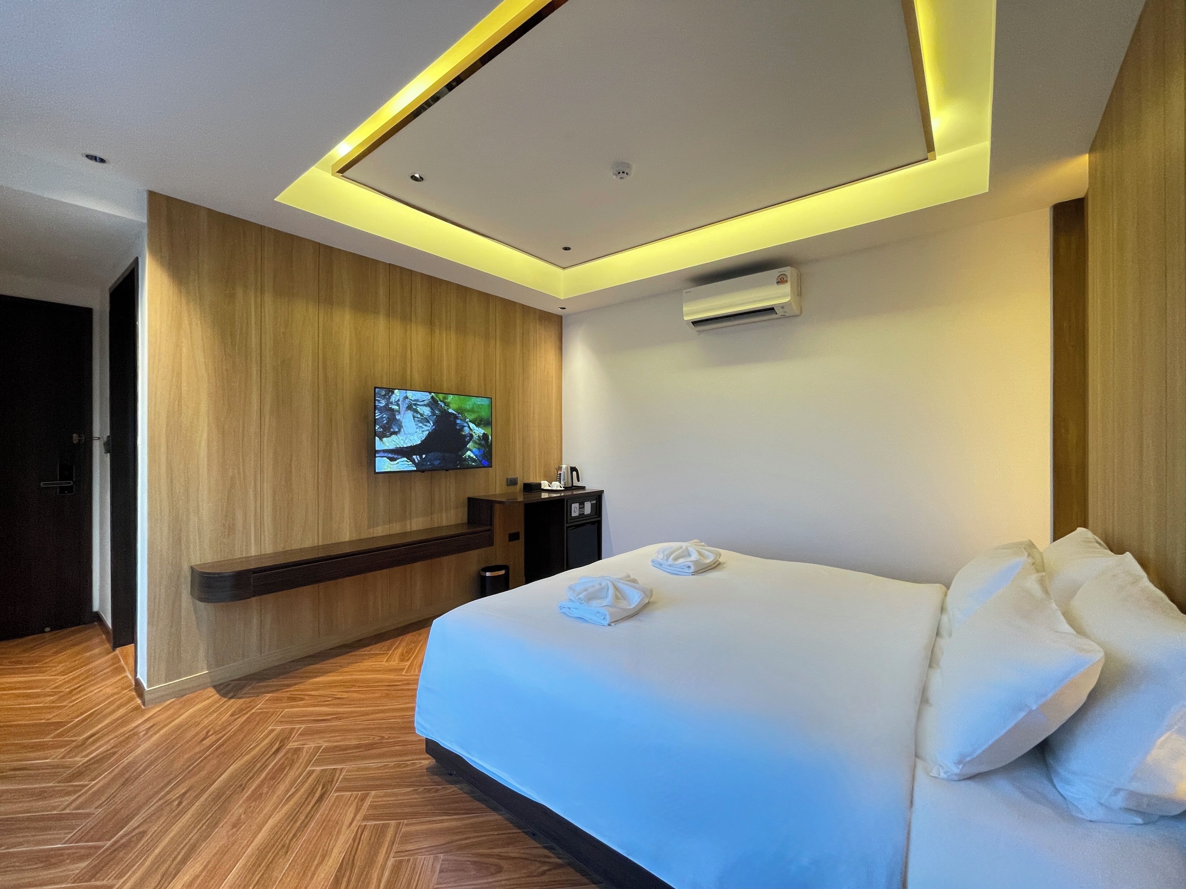 A hotel room with a large bed, a flat-screen TV mounted on the wall, a coffee machine, and an air conditioning unit.