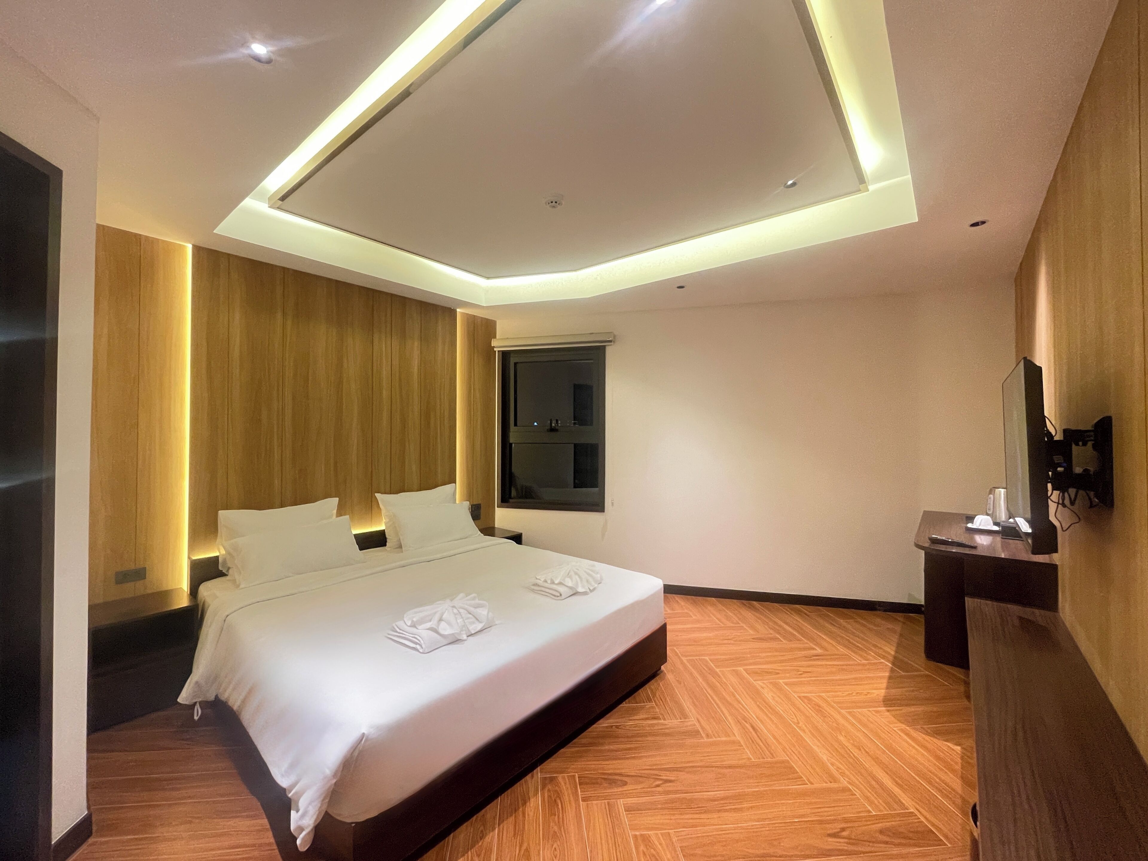 A modern hotel room with a large bed, wooden paneling, a flat-screen TV, and a desk with a computer.