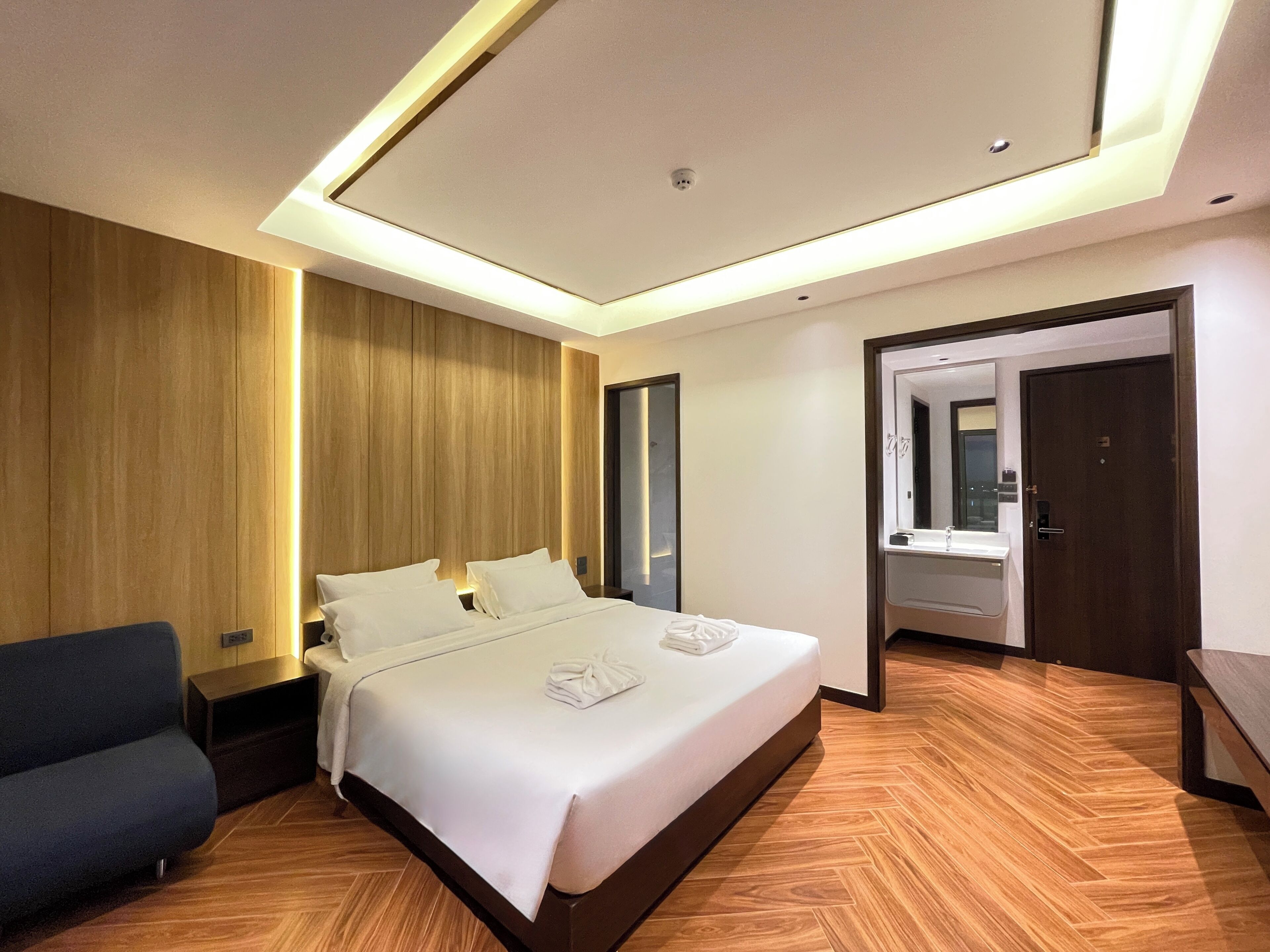 A modern hotel room with a large bed, wooden paneling on the walls, a dark blue sofa, and a bathroom with a mirror and sink.