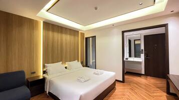 A modern hotel room with a large bed, wooden paneling on the walls, a dark blue sofa, and a bathroom with a mirror and sink.