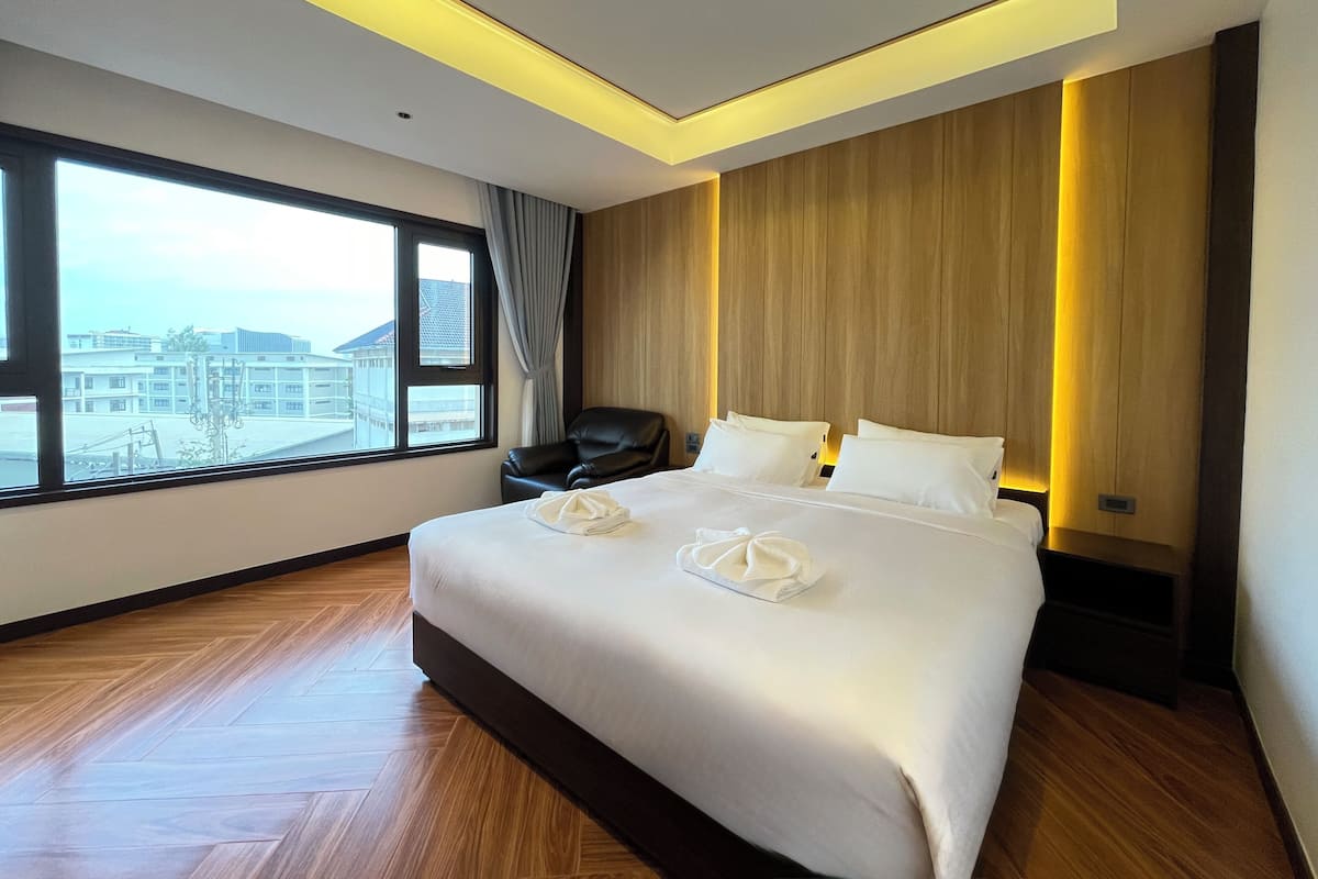 Une chambre d’hôtel moderne avec un grand lit, un plancher en bois, une vue sur les immeubles par une grande fenêtre et un fauteuil en cuir noir.