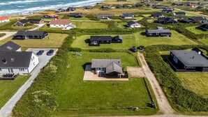 Vue aérienne d’un quartier résidentiel situé sur la côte, avec des maisons, des pelouses verdoyantes et une plage en arrière-plan.
