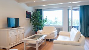 A modern living room with a white sofa, a glass coffee table, a white cabinet, a television, and a large window with curtains.