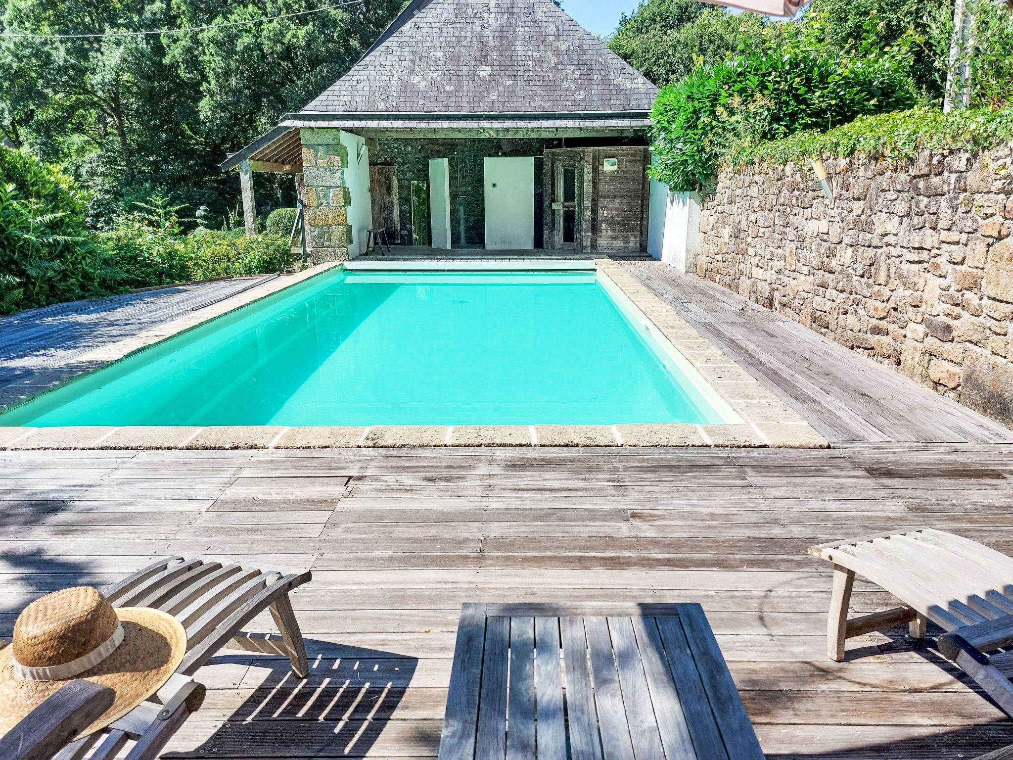 A swimming pool with a wooden deck, lounge chairs, and a stone wall.
