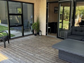 A modern outdoor deck with a wooden floor, a wicker sofa, and a glass door leading to a patio area with chairs and a table.
