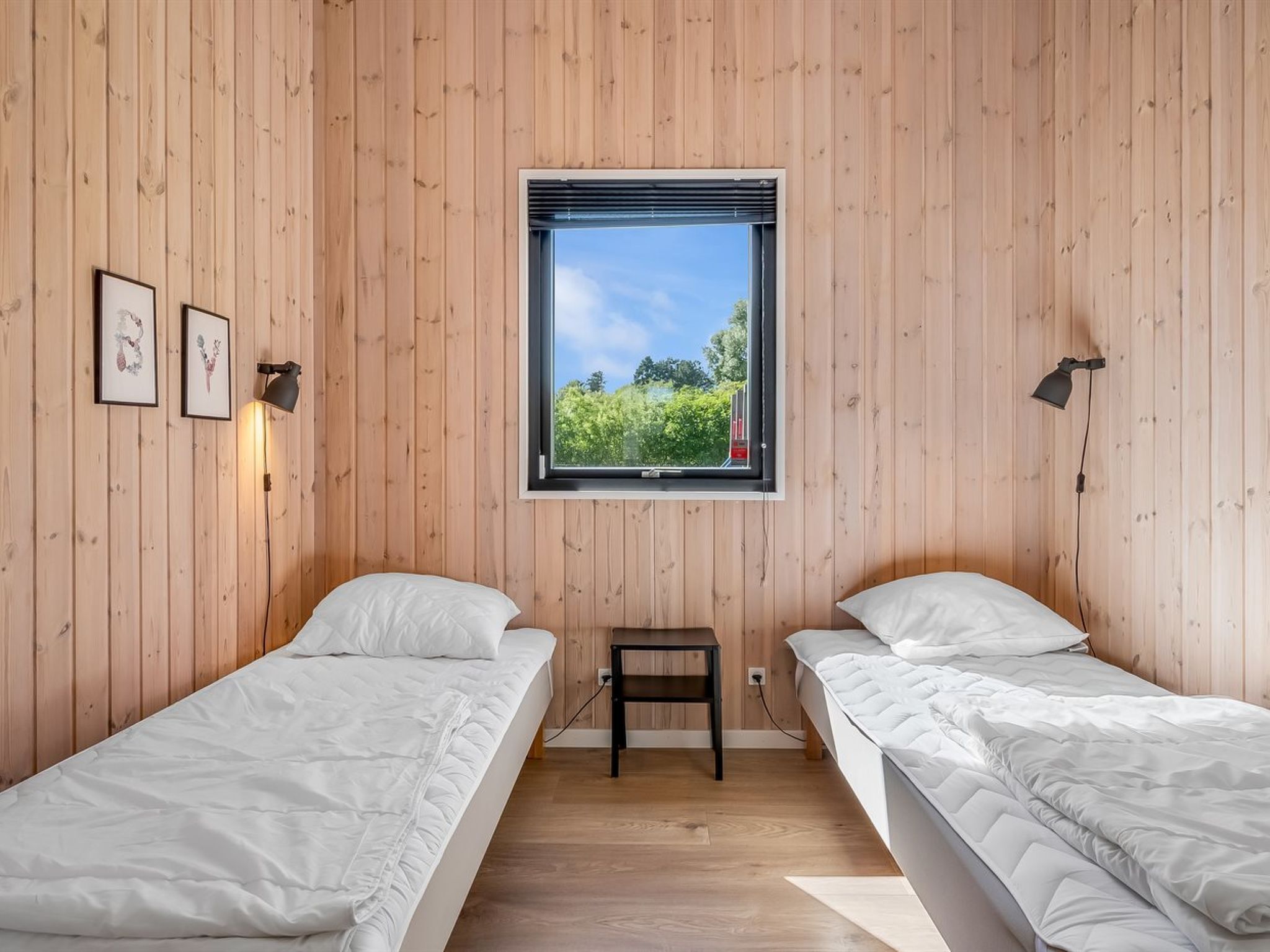 Quarto com duas camas, paredes de madeira, janela com vista para área verde e duas luminárias fixadas na parede.