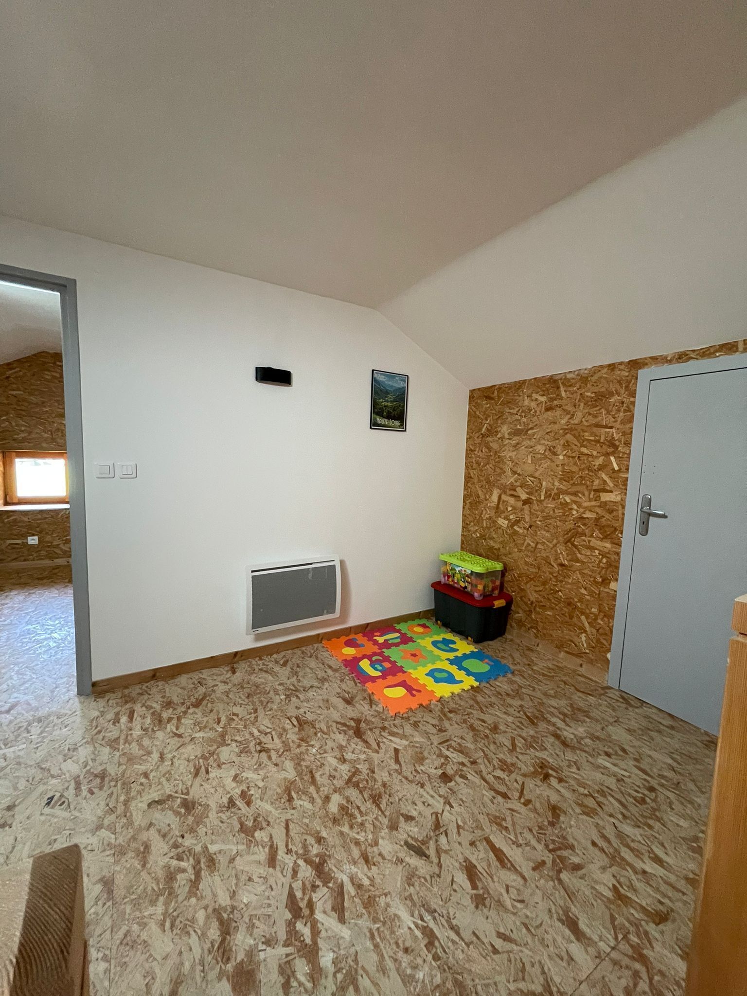 Una habitación con suelo de madera, una puerta, un respiradero pequeño, un tapete de juegos de colores y un cartel en la pared.