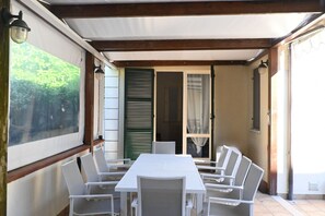 A covered patio with a table and chairs, a view of greenery, and a door with shutters.
