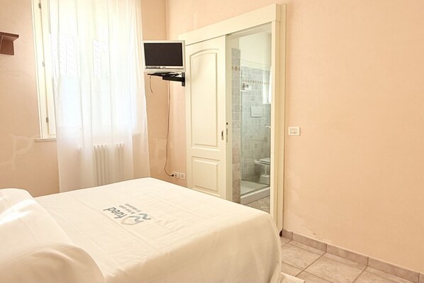 Habitación de hotel con cama, televisor montado en la pared, baño con cabina de ducha de vidrio y puerta que da a otra habitación.