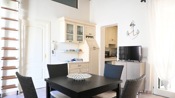 Uma sala de jantar com mesa, cadeiras, uma televisão e uma cozinha ao fundo.