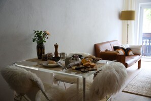 A dining table with a glass top, surrounded by white furry chairs. A vase with flowers and a pepper grinder are on the table. A brown leather sofa is in the background.