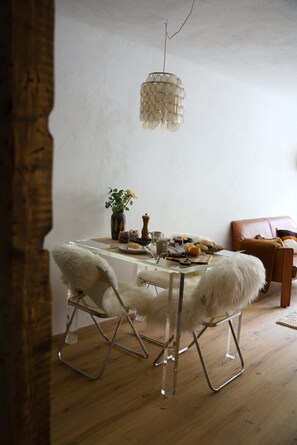 A dining area with a clear glass table, white furry chairs, a hanging light fixture, and a wooden floor.