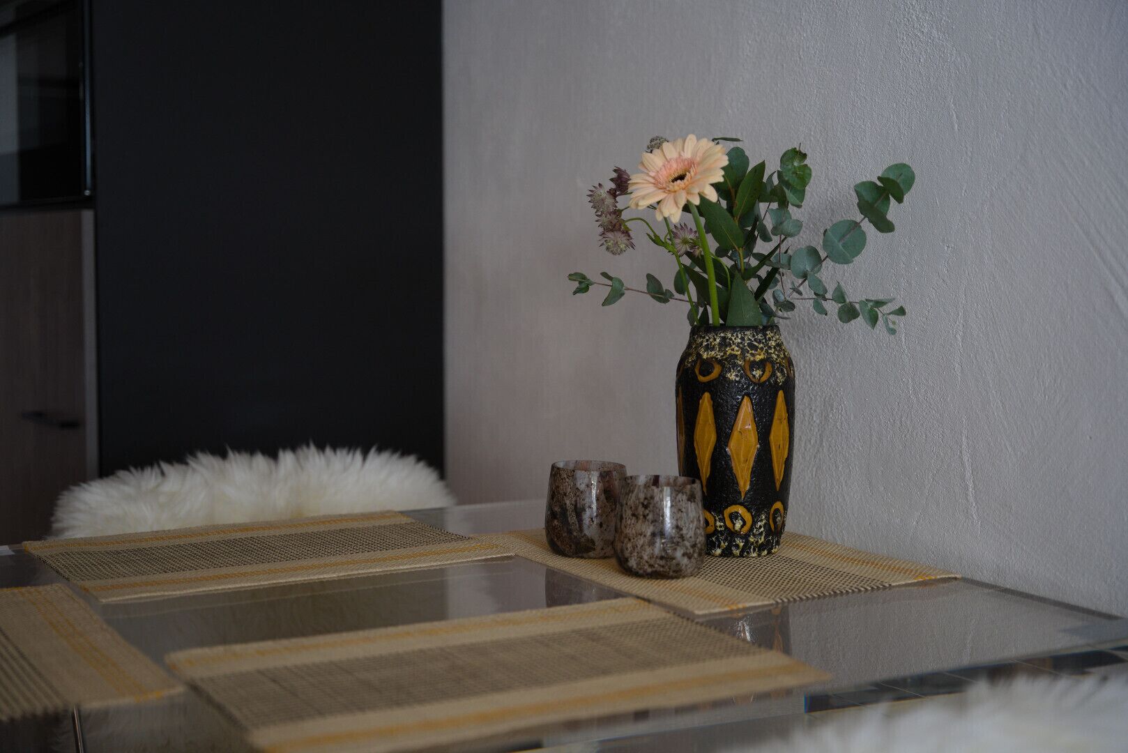 A table with a vase of flowers, a fluffy white pillow, and a woven placemat.
