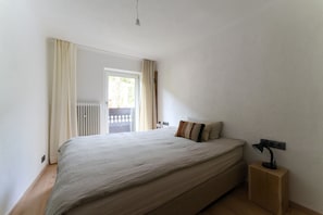 A bedroom with a large bed, a bedside table with a lamp, a window with curtains, and a view of the outside.