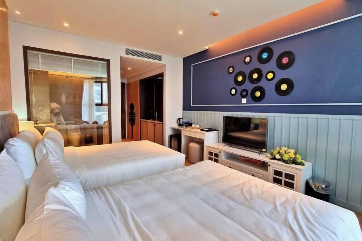 A hotel room with two beds, a TV, a wall with vinyl records, and a bathroom visible through a glass door.