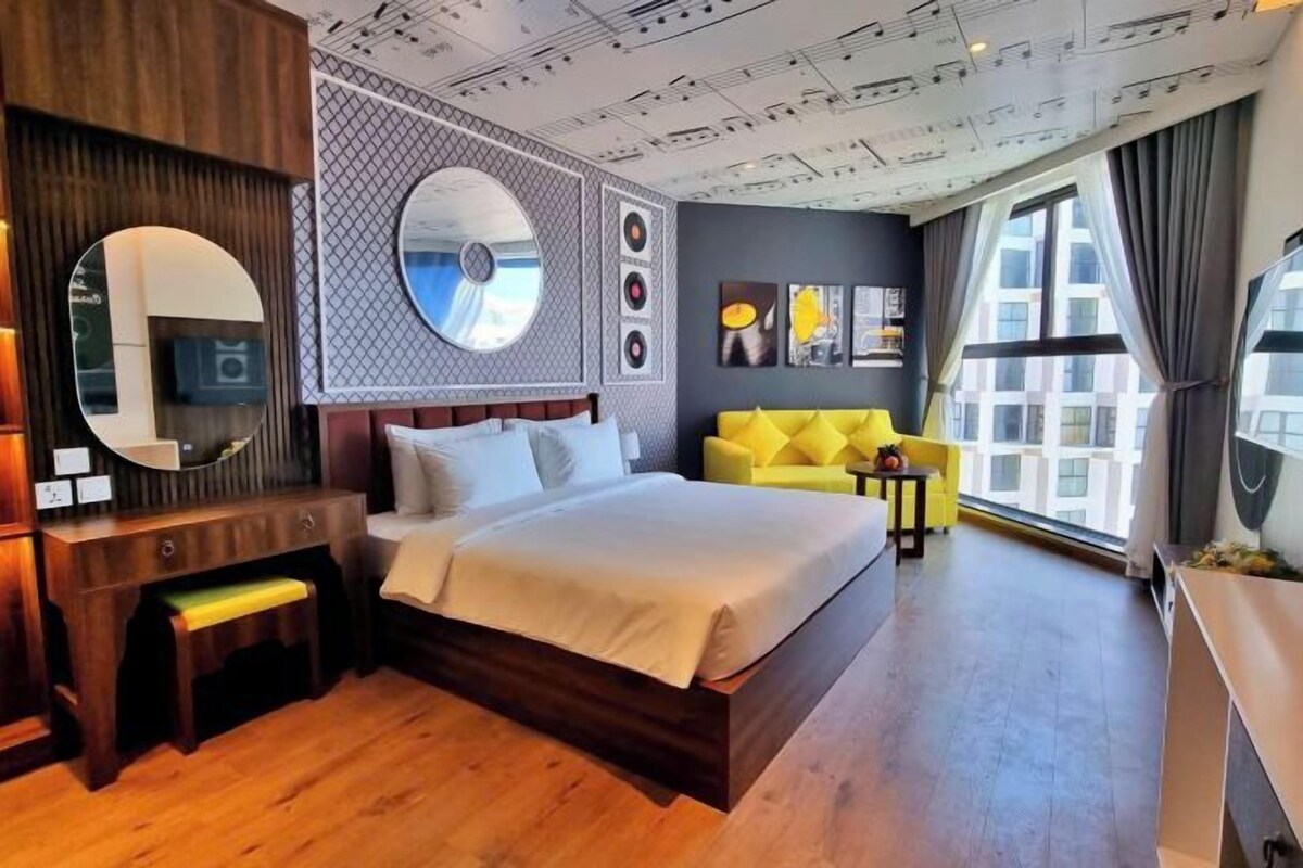 Una habitación de hotel moderna con una cama grande, un escritorio de madera, un sofá amarillo y un ventanal con cortinas.